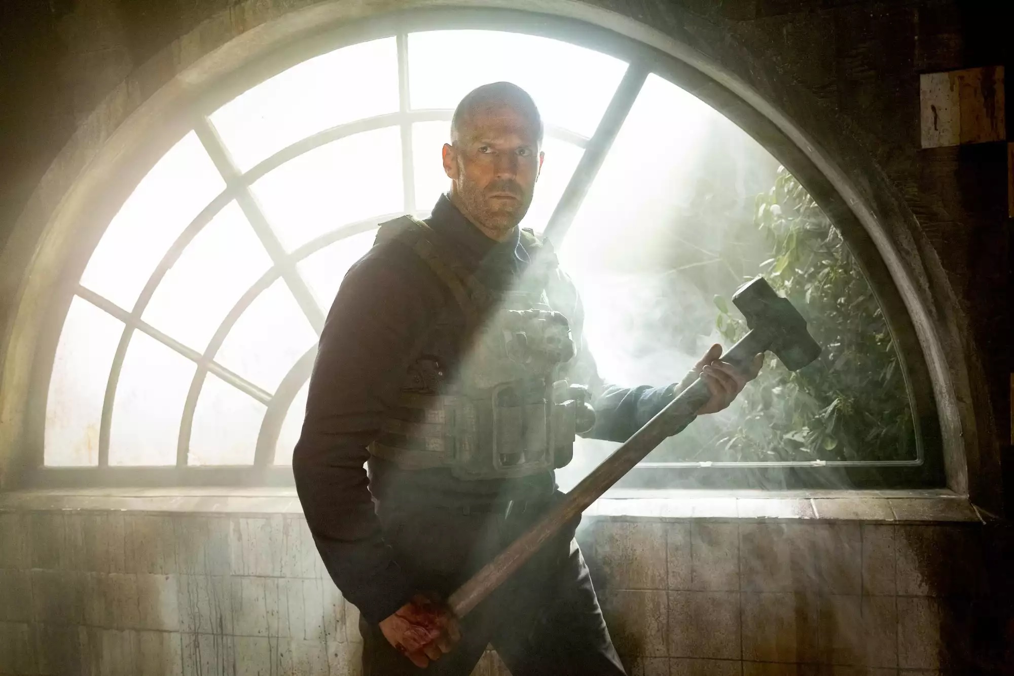 Jason Statham fica em um raio de sol na frente de uma grande janela de meia-lua e levanta uma marreta em um homem que trabalha
