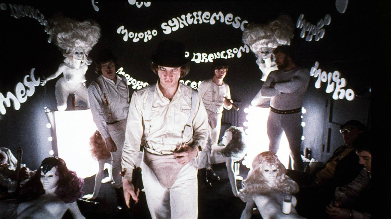 Malcolm McDowell como Alex em um relógio laranja fica em uma barra de leite escurecida cheia de manequins femininas nuas, com nomes de drogas fictícios por todas as paredes