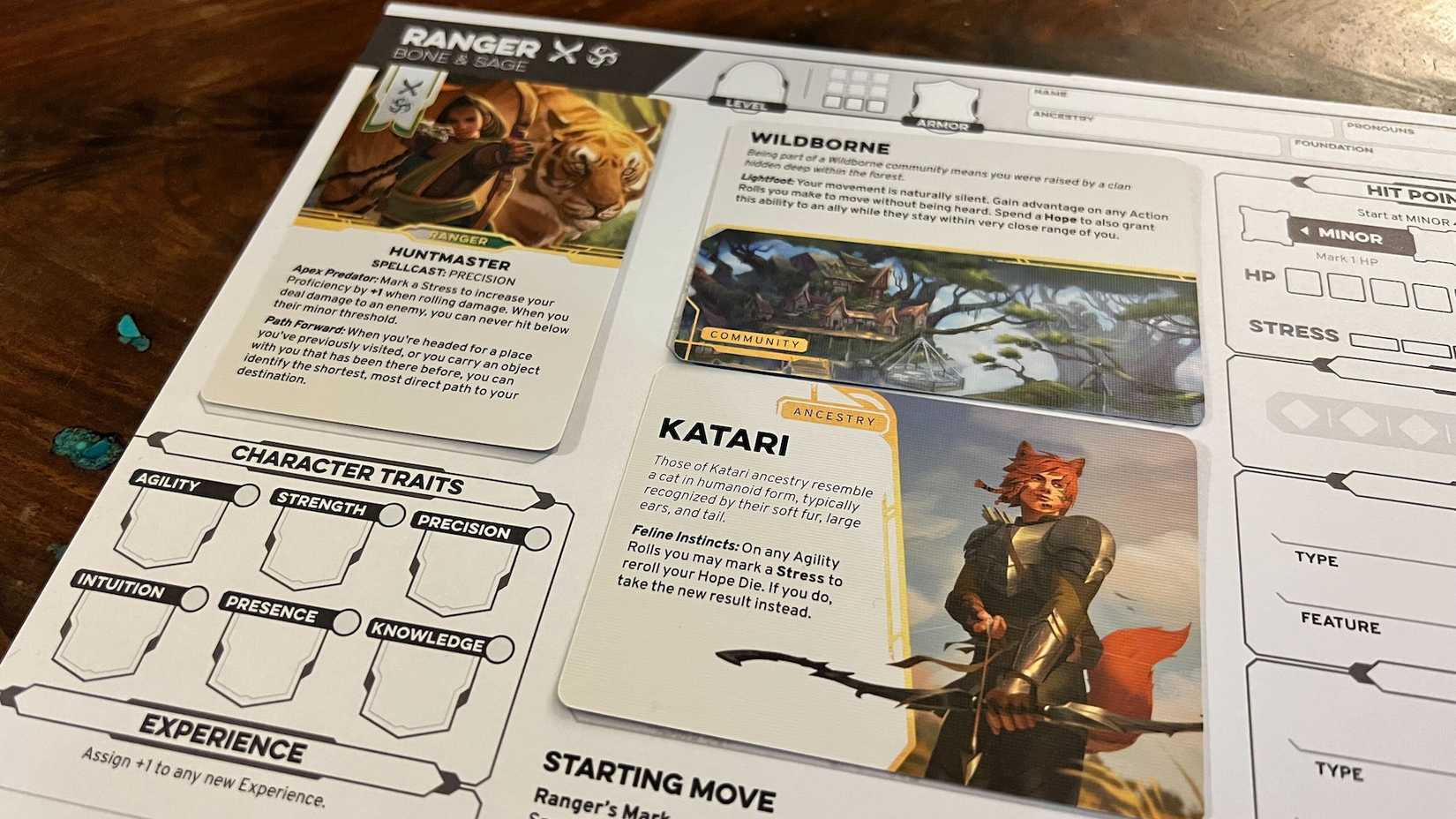 Um close da folha de personagens baseada em cartão em Daggerheart. Um cartão, para Huntmaster, chama a aula. Outro, para Katari, chama a ascendência do caráter, enquanto outra - com base selvagem - observa a comunidade à qual pertence.