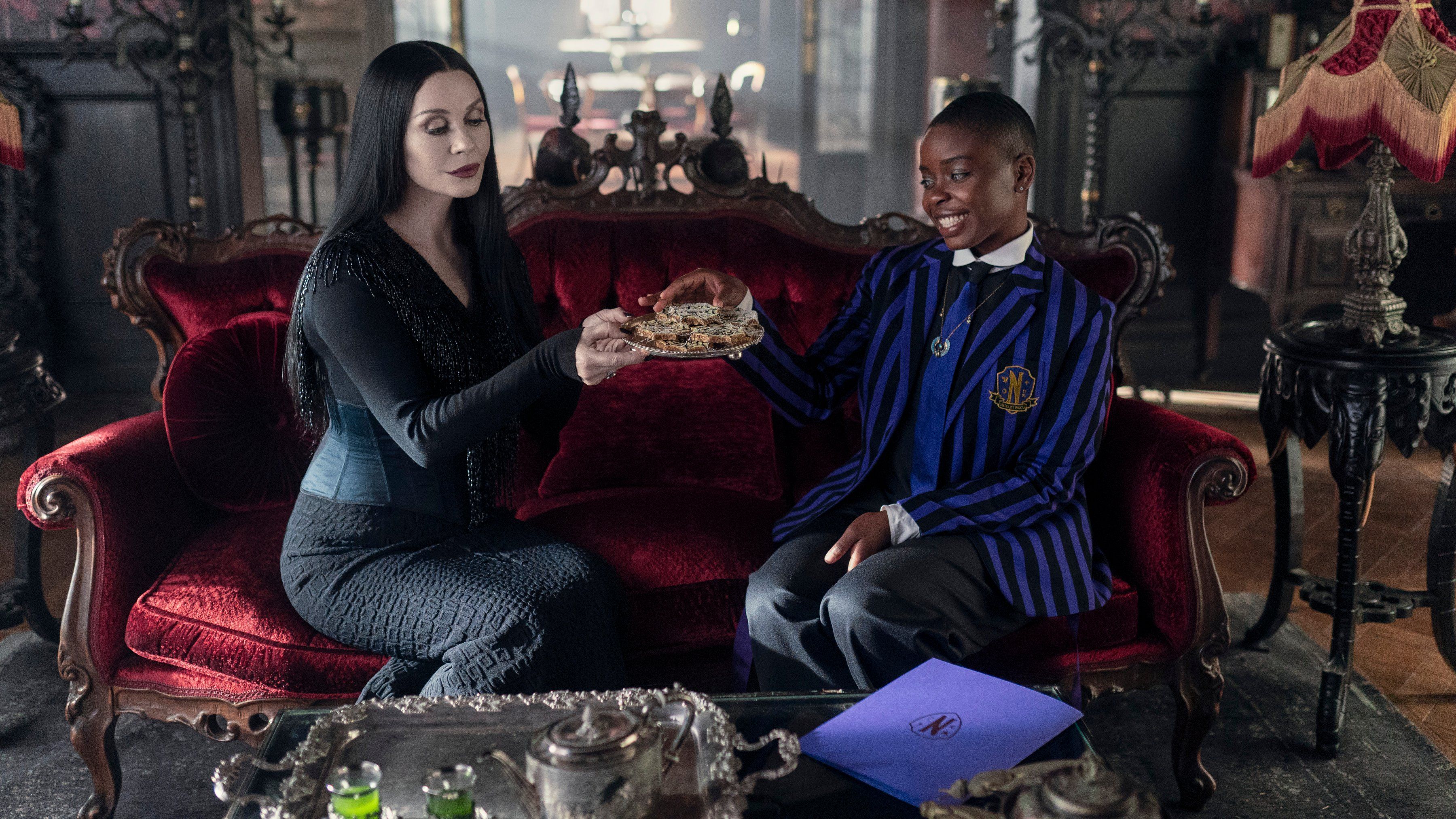 Catherine Zeta-Jones como Morticia Addams mantém uma bandeja de biscoitos enquanto está sentado em um sofá de alegria no domingo como Bianca Barclay na quarta-feira