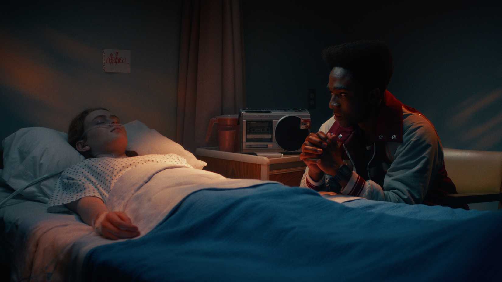 Max Mayfield (Sadie Sink) deita -se em uma cama de hospital com Lucas Sinclair (Caleb McLaughlin) sentado ao seu lado