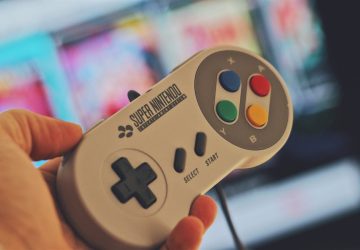 Hand holding a classic Super Nintendo game controller indoors with a blurred screen in the background.