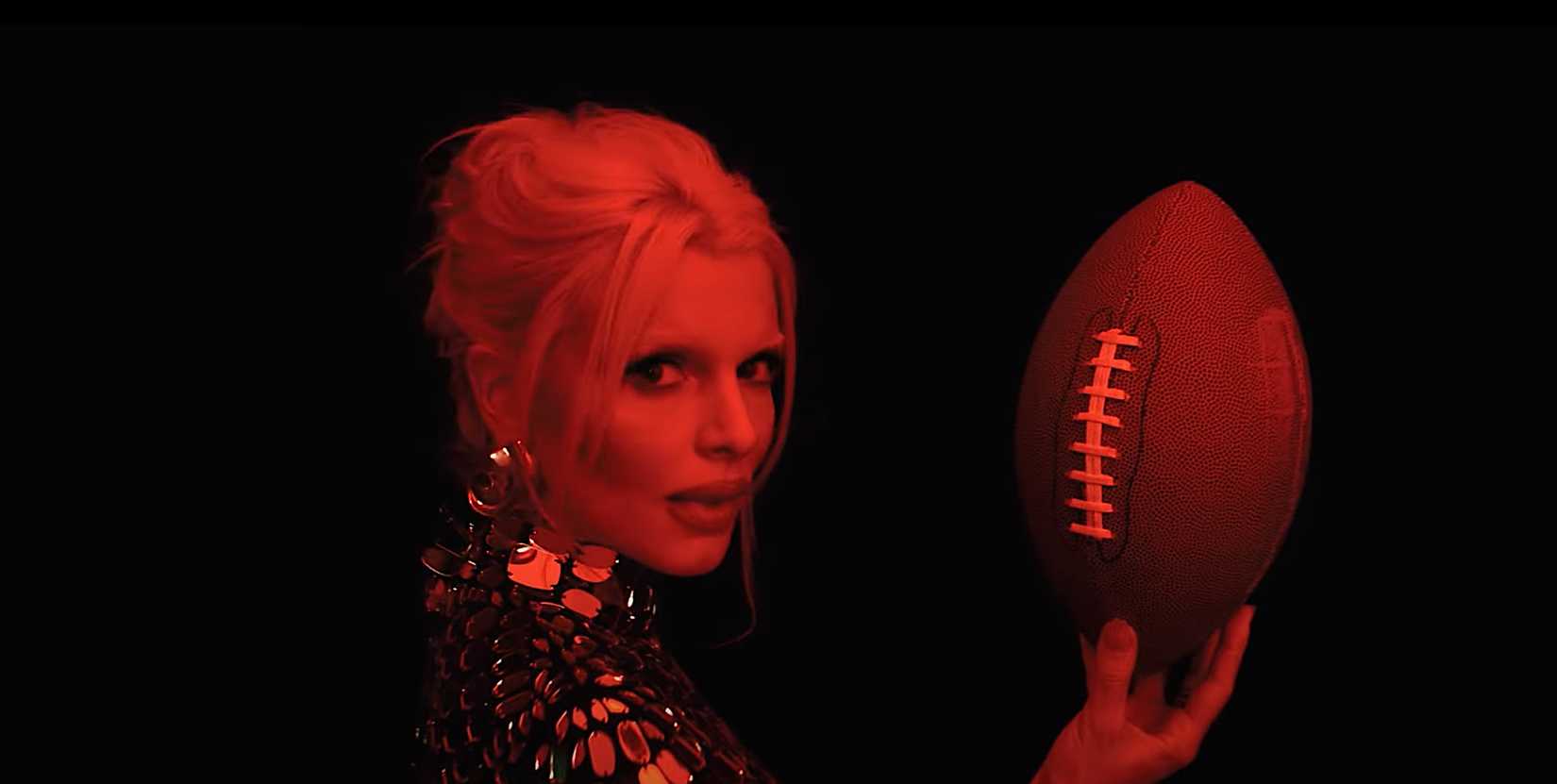 Julia Fox nele, uma mulher em um vestido brilhante com o cabelo puxado para trás, segurando uma bola de futebol, iluminada pela luz vermelha de sangue