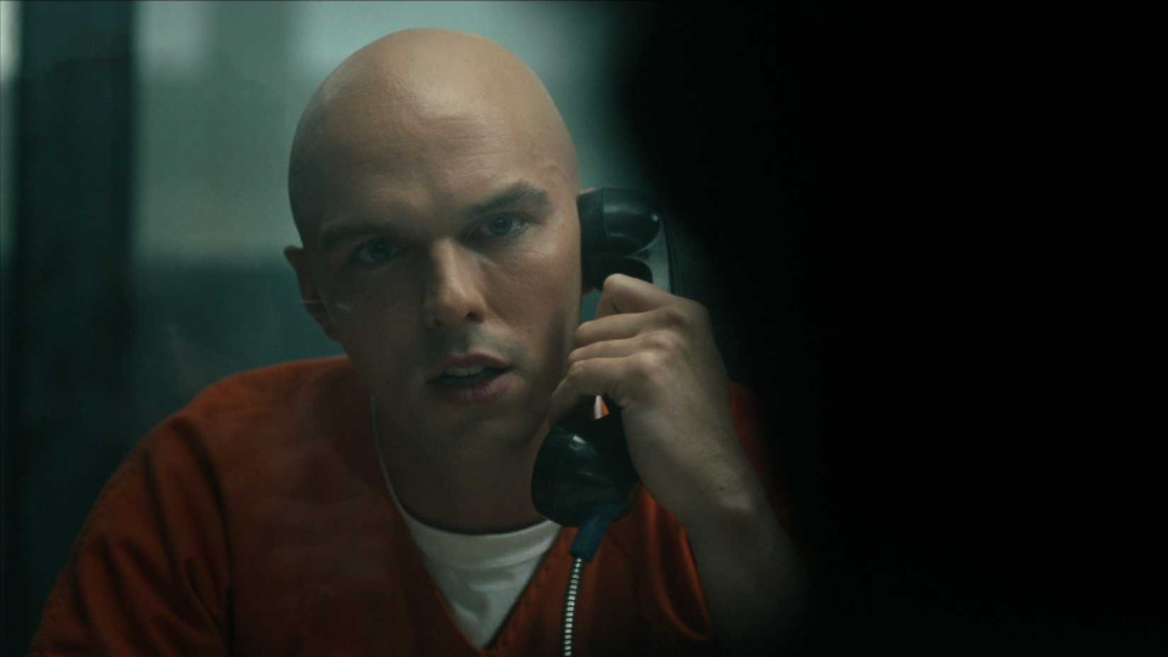 Nicholas Hoult como Lex Luthor, na prisão laranja, fala com uma figura sombria da tela em pacote em pacificador