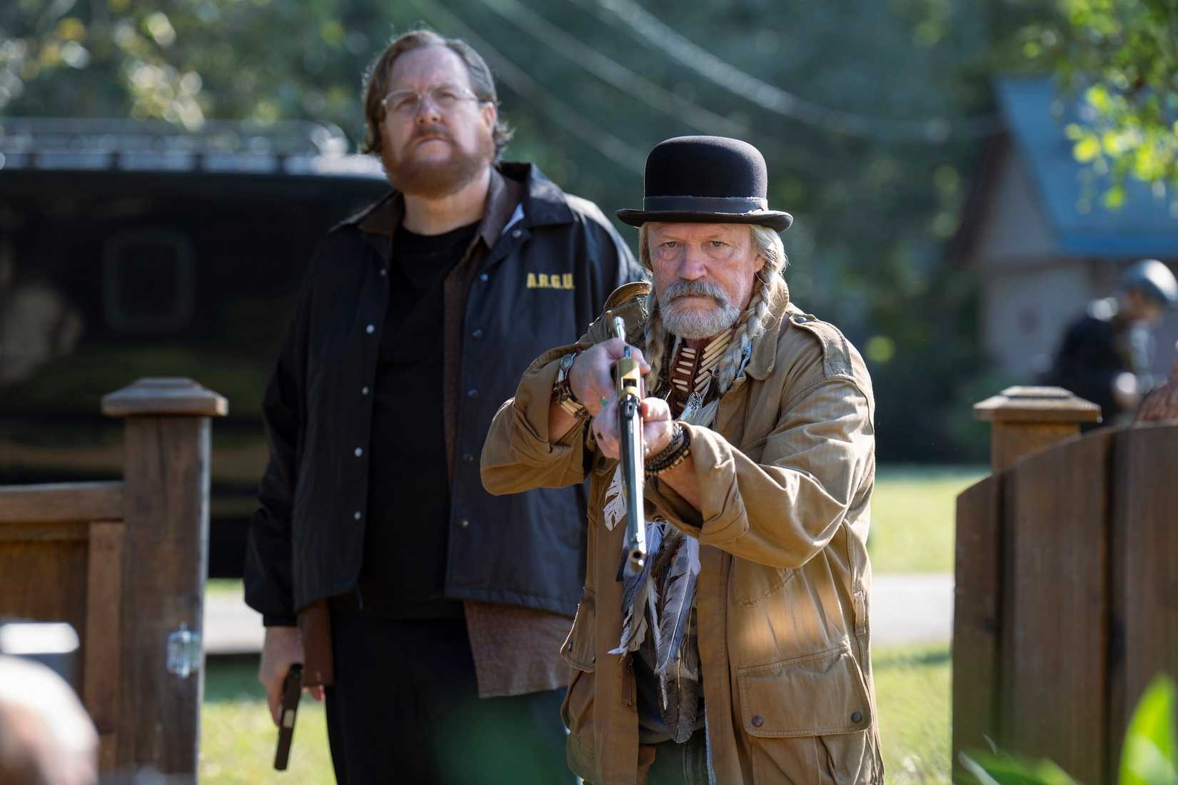 Michael Rooker com duas tranças longas e um espanador que segura um rilfe com um grande cara atrás dele no pavão da segunda temporada do episódio 4