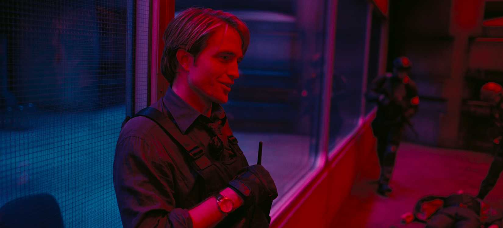 Robert Pattinson usando um colete à prova de balas como Neil em princípios lavados com luz vermelha e azul