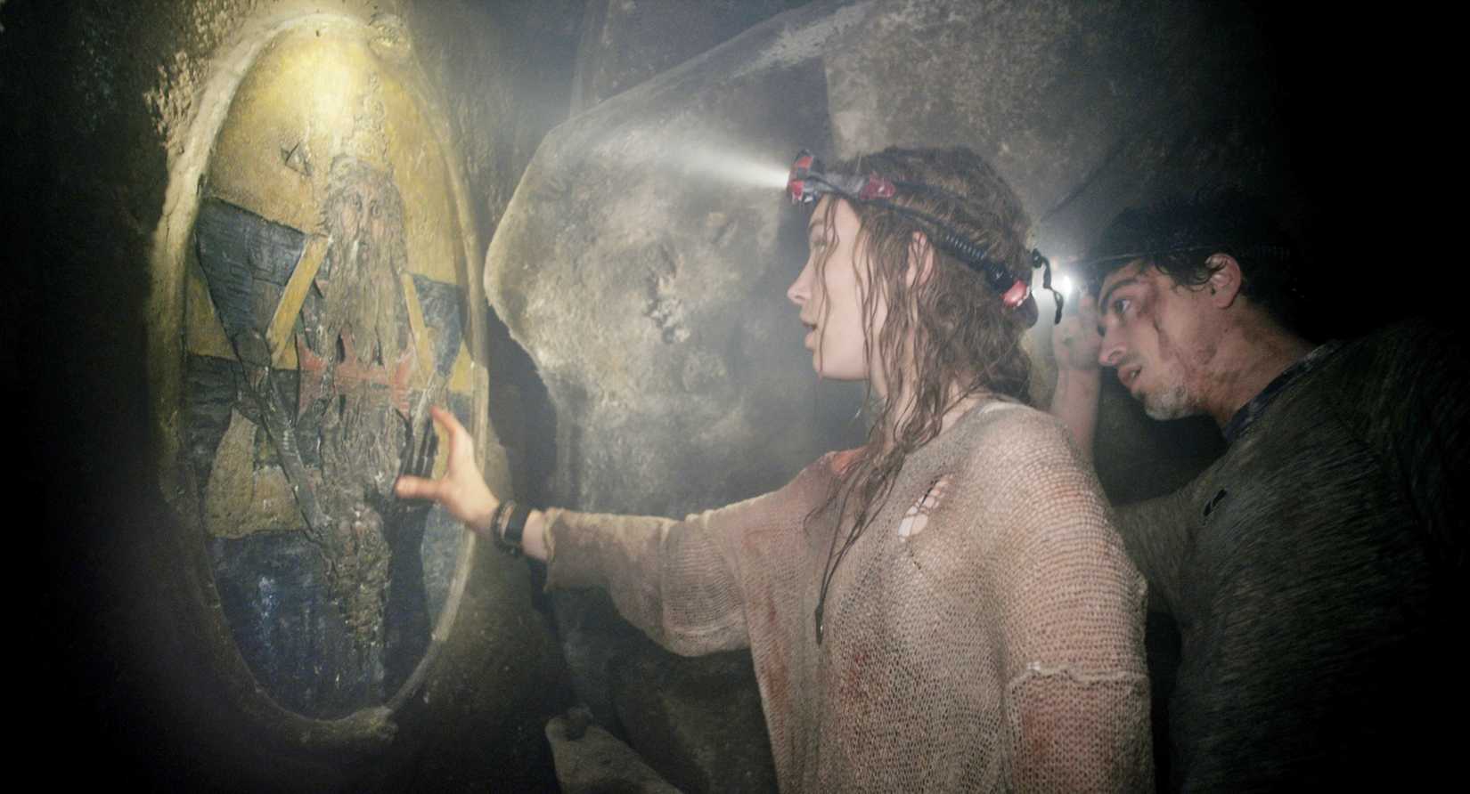 Duas pessoas desgrenhadas e manchadas de sangue (Perdita Weeks e Ben Feldman) apontam lanternas para um antigo símbolo oculto em uma parede em As Above, So Below