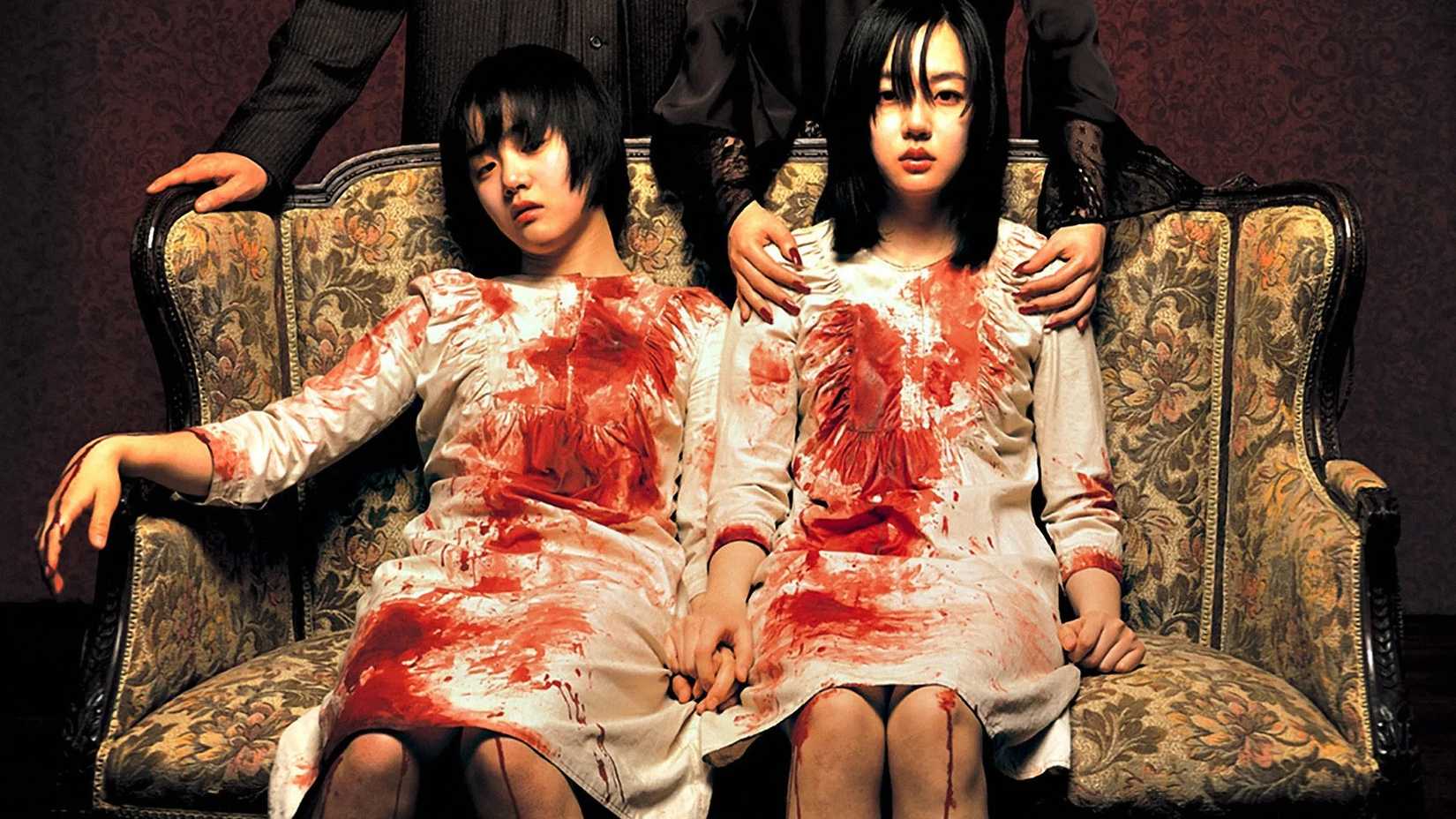Duas mulheres coreanas com vestidos brancos cobertos de sangue estão sentadas lado a lado em um sofá de brocado, uma delas com uma figura invisível parada atrás e acima dela, com as mãos firmemente nos ombros, em A Tale of Two Sisters