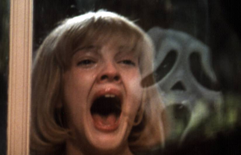 Drew Barrymore está em frente a uma janela, gritando de medo, enquanto o assassino mascarado Ghostface é refletido no vidro em Pânico, de 1996.