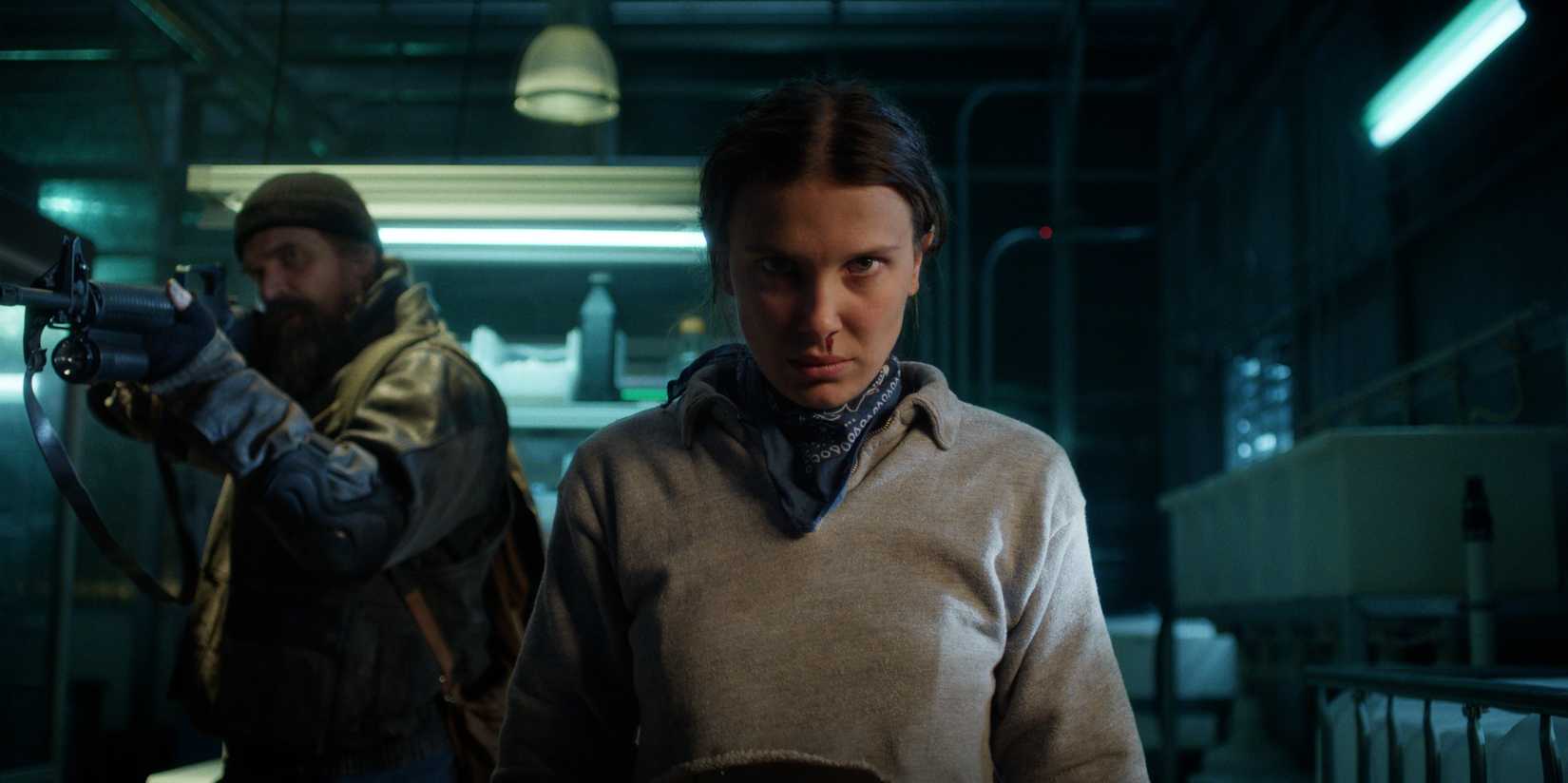 David Harbor como Jim Hopper segurando uma espingarda está atrás e Millie Bobby Brown como Eleven com sangramento nasal na 5ª temporada de Stranger Things