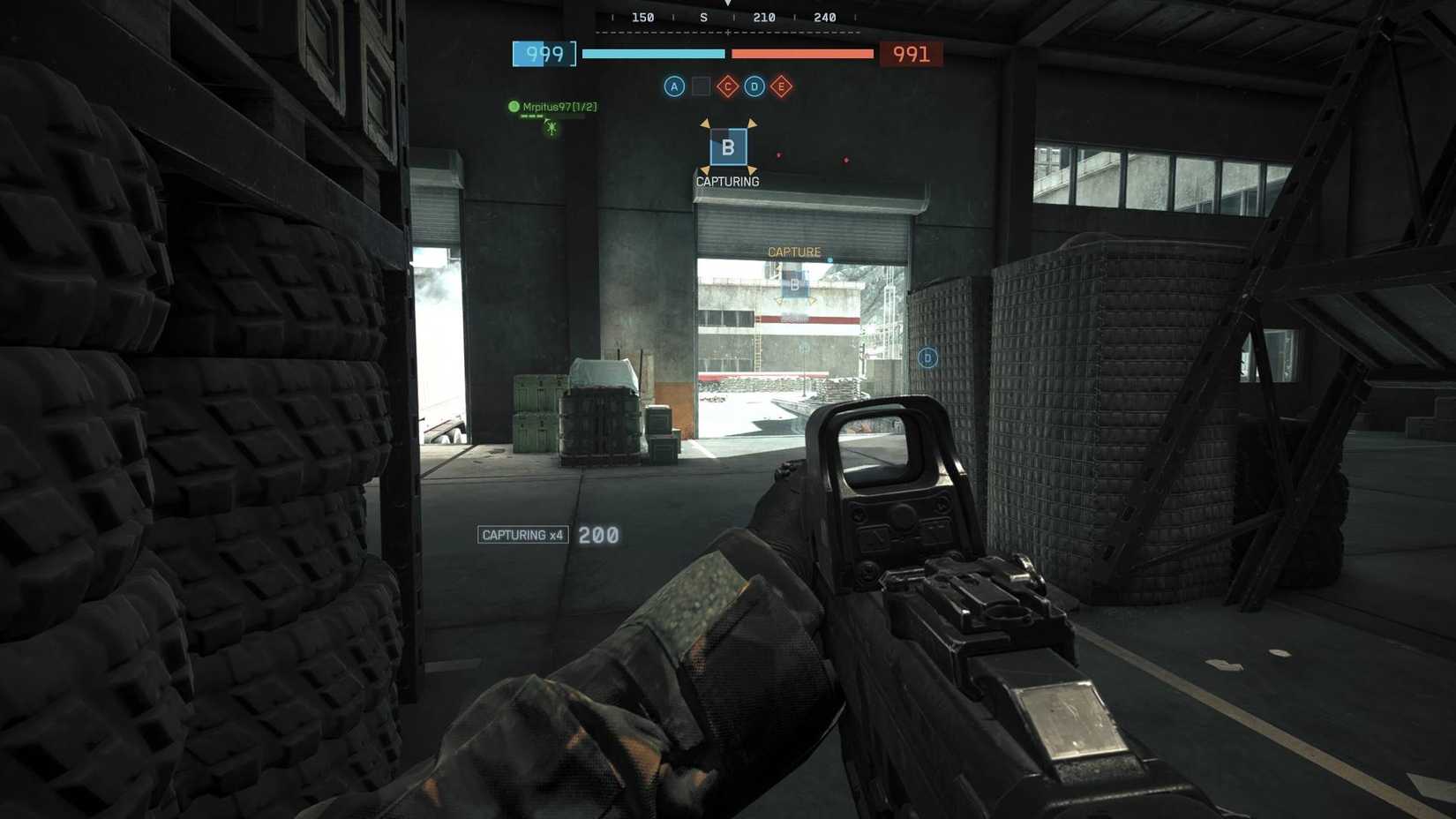 O jogador que captura B em Conquest na Operação Firestorm, equipado com um rifle de assalto escondido em um canto de um hangar.