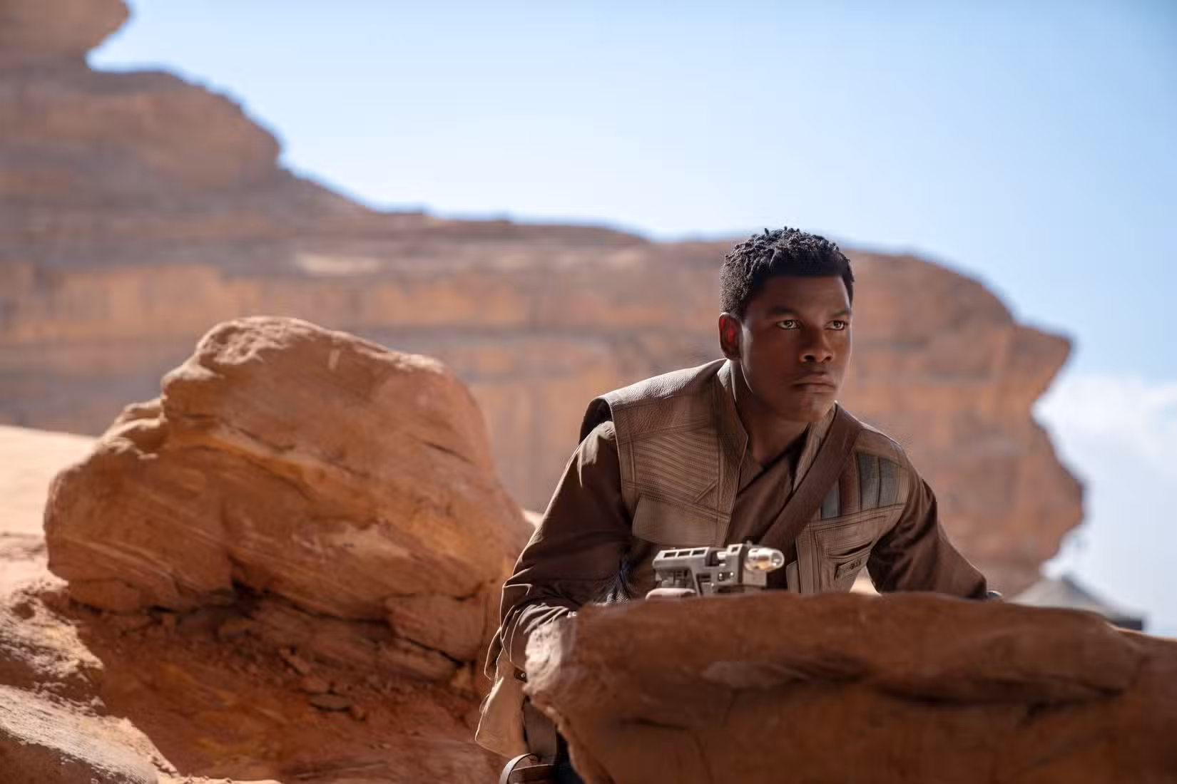 Imagem de John Boyega como Finn em The Rise of Skywalker. Ele está cercado por um local deserto e se esconde entre as rochas.