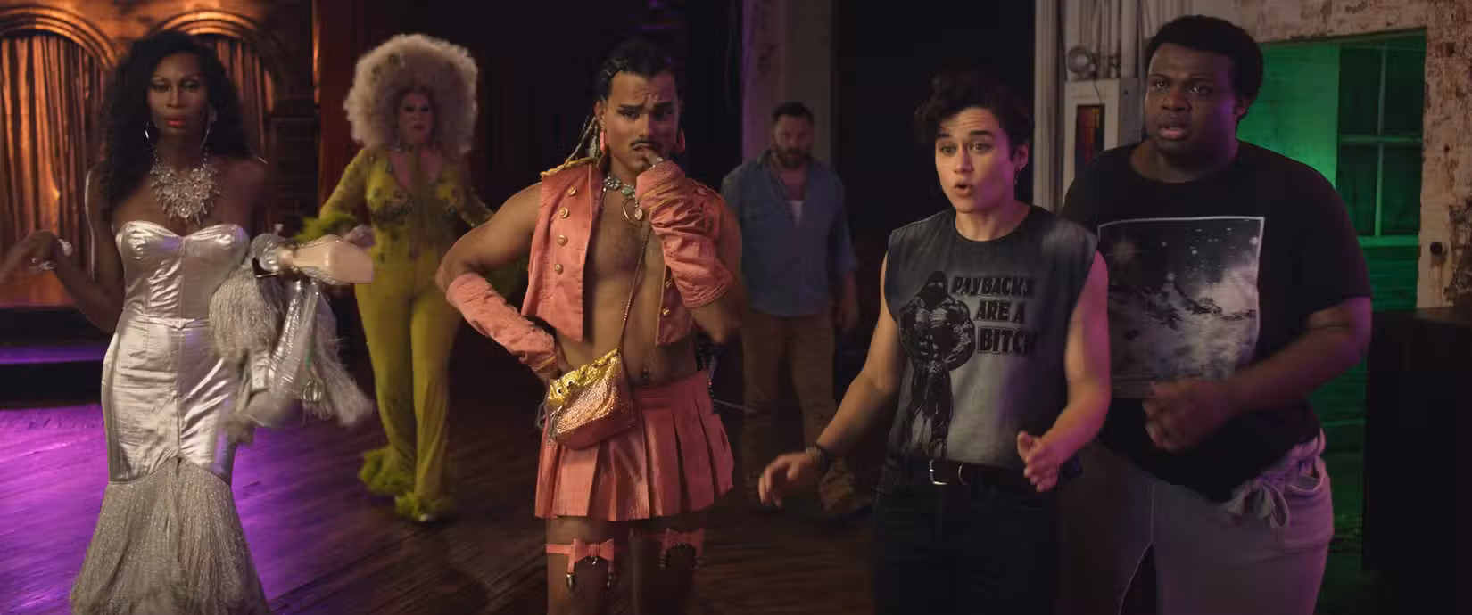 Eu escolhi isso: um grupo de personagens de Queens of the Dead, todos vestidos com diferentes formas de fabuloso (um vestido de baile sem mangas em lamê prateado, uma enorme peruca branca fofa e meia amarelo mostarda, um colete aberto sem camisa por baixo, uma camisa masculina sem mangas que diz "As vinganças são uma merda," e mais) ficam juntos, parecendo preocupados ou alarmados