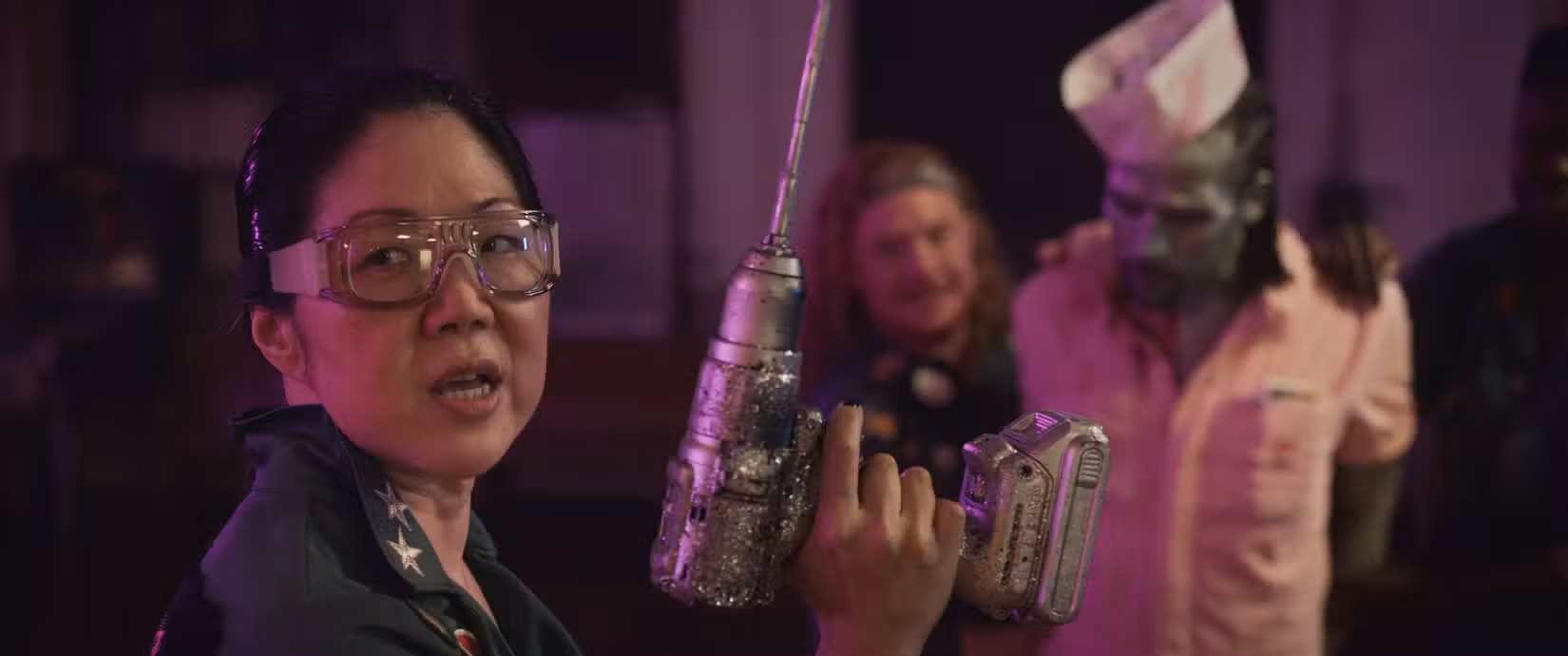 Margaret Cho segura uma broca de prata enquanto um zumbi verde surge atrás dela em Queens of the Dead, de Tina Romero.