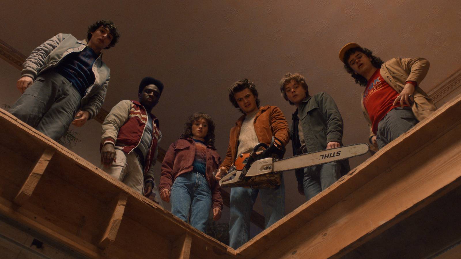 Finn Wolfhard como Mike Wheeler, Caleb McLaughlin como Lucas Sinclair, Natalia Dyer como Nancy Wheeler, Joe Keery como Steve Harrington, Charlie Heaton como Jonathan Byers e Gaten Matarazzo como Dustin Henderson estão na frente de um buraco em um prédio enquanto Steve segura uma serra elétrica em Stranger Things
