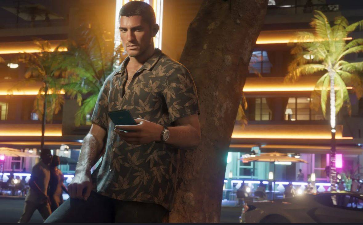 Um dos dois protagonistas no GTA 6, Jason, segura um telefone enquanto ele se inclina contra uma árvore. Ele está usando uma camisa estampada com folhas enquanto a vida noturna se agita ao fundo. 