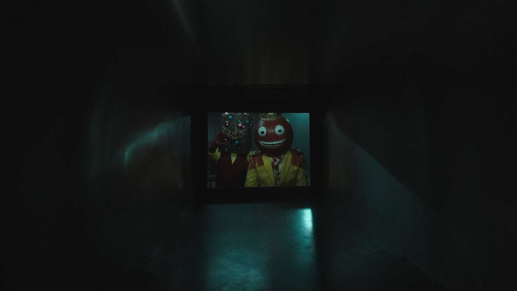 Duas criaturas assustadoras-uma em um terno amarelo, com um gumbal sorridente para uma cabeça e o outro com uma cabeça de barra de doces-aparecem em uma pequena janela no final de um longo túnel de metal no Halloween V/H/S "Tamanho divertido"