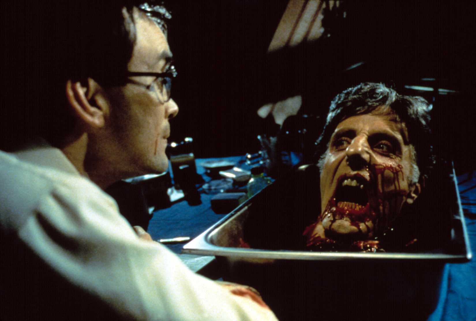 O cientista revestido de laboratório (?) Herbert West (Jeffrey Combs) fica calmamente como uma cabeça coberta de sangue, cortada e reanimada, grita com ele de uma bacia em seu balcão de laboratório no re-animador