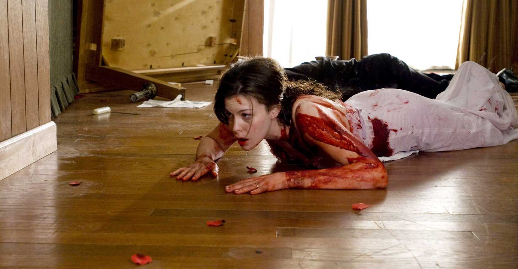 Uma mulher ensanguentada com um vestido claro (Liv Tyler) rasteja pelo chão de madeira no filme The Strangers, de 2008