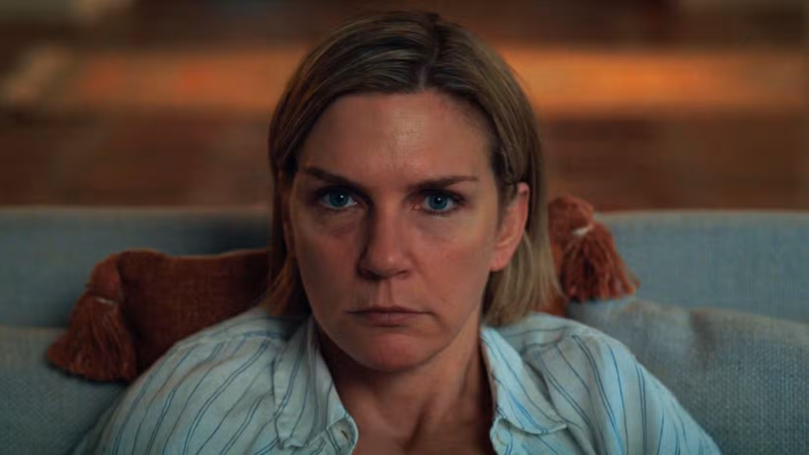 Carol (Rhea Seehorn) olha diretamente para frente enquanto a TV passa. Ela está no sofá, com uma almofada atrás dela.