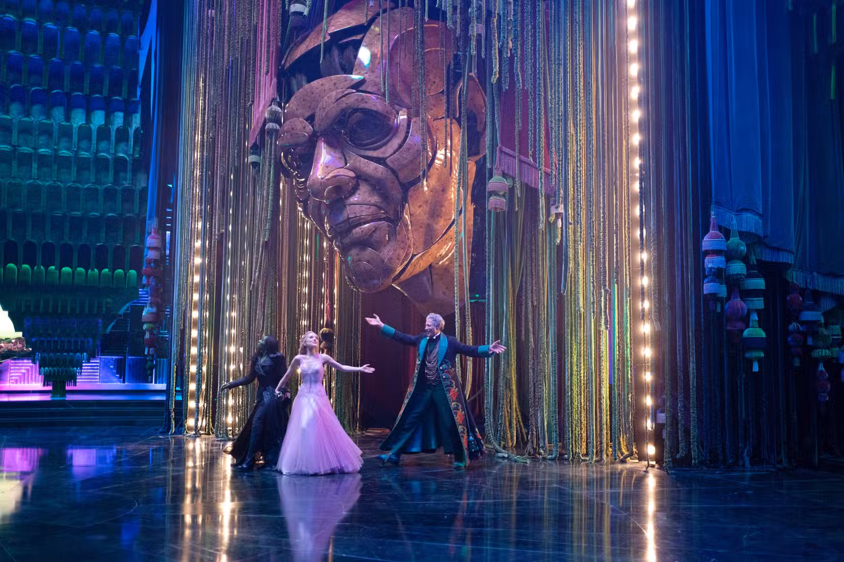 Elphaba (Cynthia Erivo), Glinda (Ariana Grande) e o Mágico de Oz (Jeff Goldblum) dançam juntos em sua câmara de audiência, ofuscados pela enorme cabeça artificial do Mágico de Oz, em Wicked: For Good