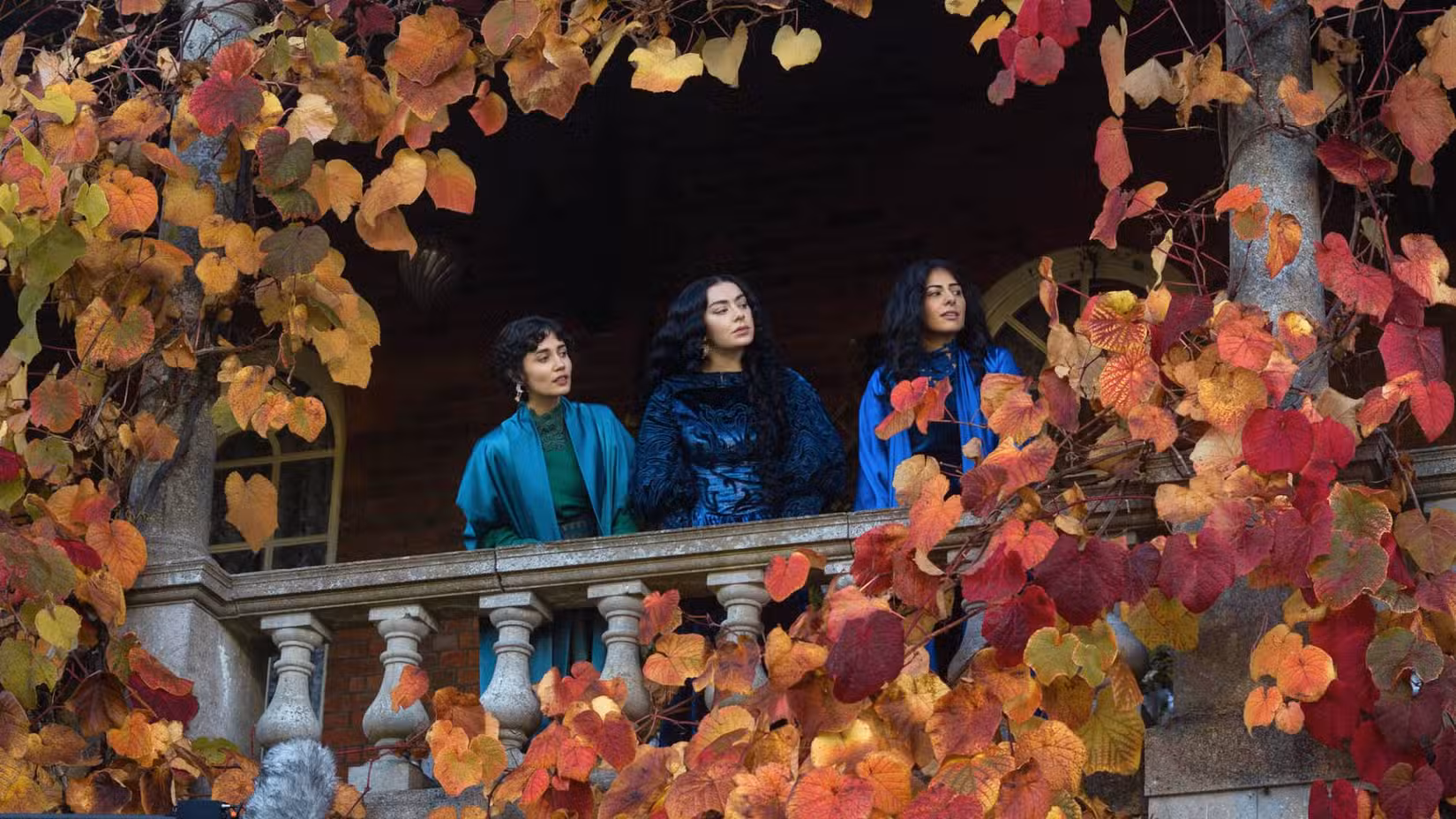 Uma imagem de 100 Noites de Herói. Mostra três mulheres em uma varanda, cercadas por folhas e outras folhagens. As três mulheres usam lindos vestidos azuis.