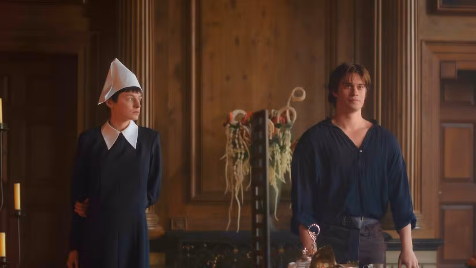 Emma Corrin e Nicholas Galitzine de 100 Noites de Herói. Os dois ficam lado a lado, com Corrin em um vestido preto com gola branca. Galitzine veste uma camisa preta, com gola larga para acentuar o peito. 