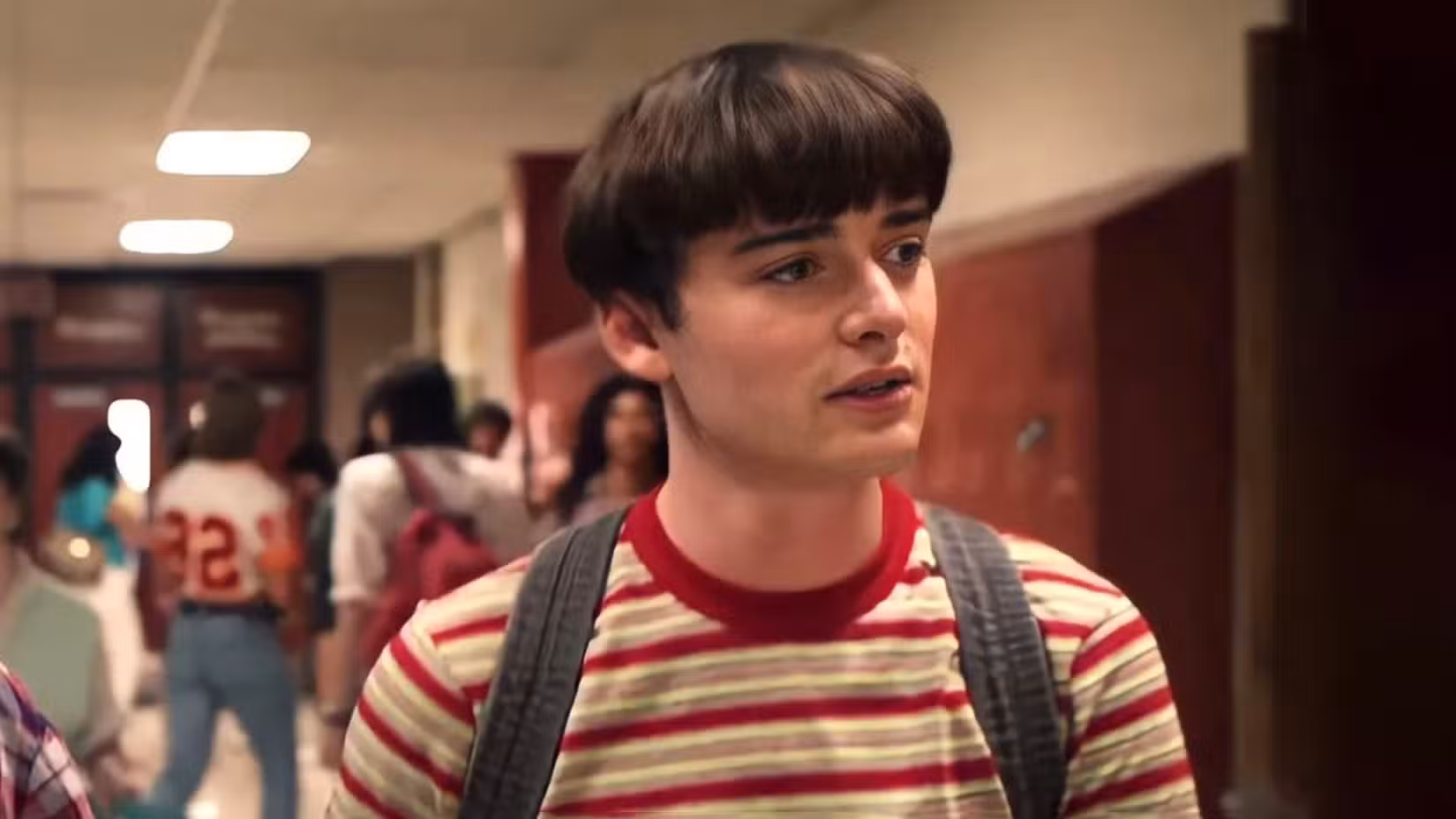 Noah Schnapp como Will Byers, andando pelo corredor do colégio com um corte tigela e uma camisa listrada vermelha e amarela enquanto usava uma mochila em Stranger Things