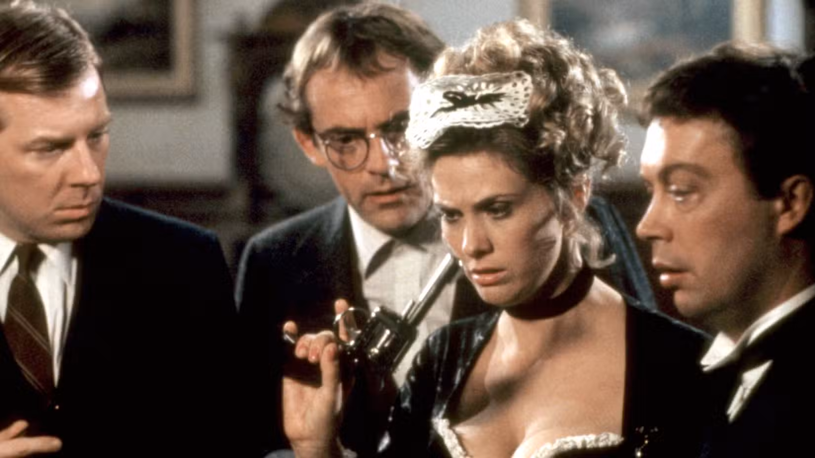 A empregada francesa Yvette (Colleen Camp) segura uma arma enquanto está cercada pelo Sr. Green (Michael McKean), Professor Plum (Christopher Lloyd) e Wadsworth (Tim Curry)