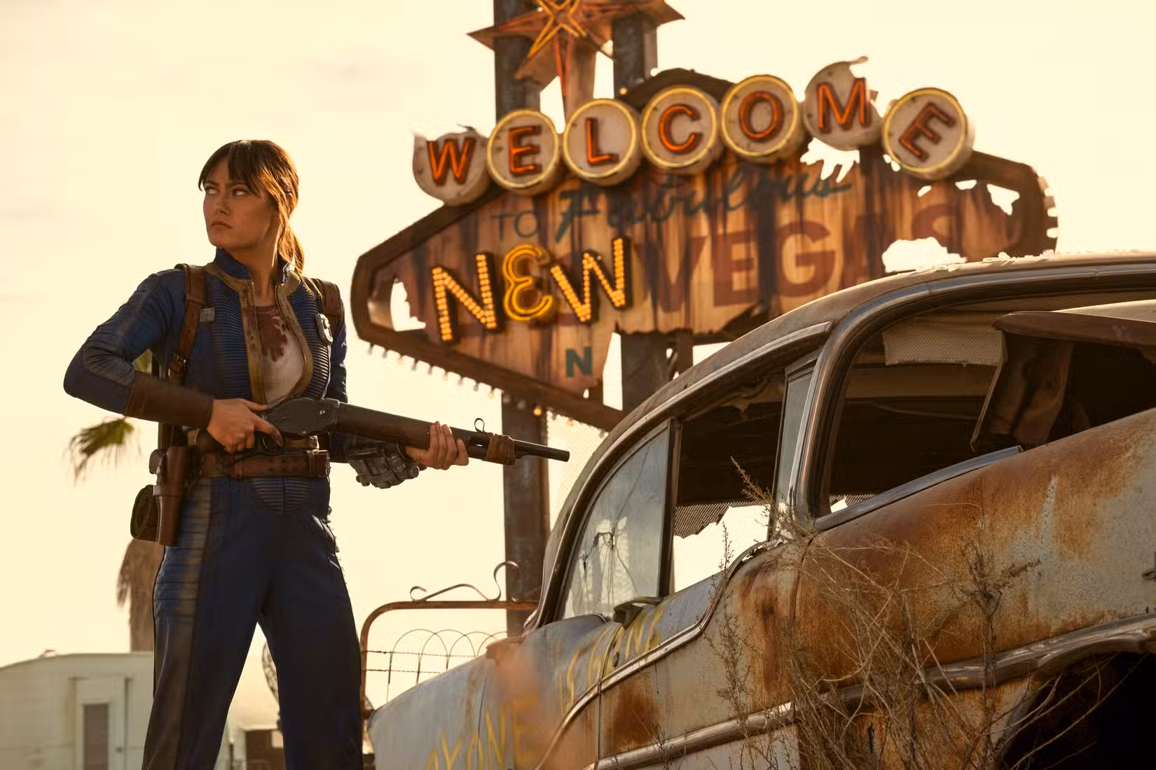 Lucy (Ella Purnell) está parada, posando com uma espingarda, em frente à placa de Bem-vindo ao novo Vegas na 2ª temporada de Fallout.