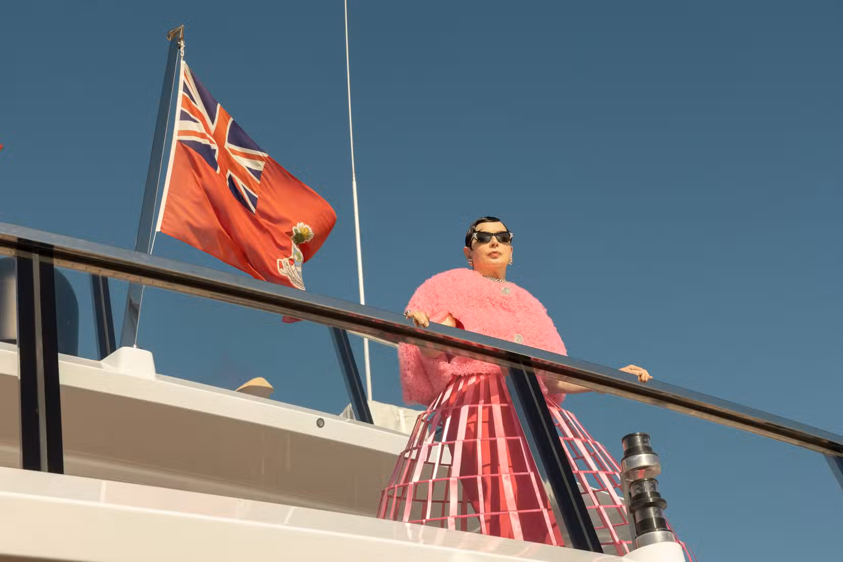 Uma imagem de A Bela (2026). Ele mostra uma mulher com uma jaqueta rosa fofa, óculos escuros e uma saia de arame que vai até o chão, encostada na grade do convés superior de um iate.
