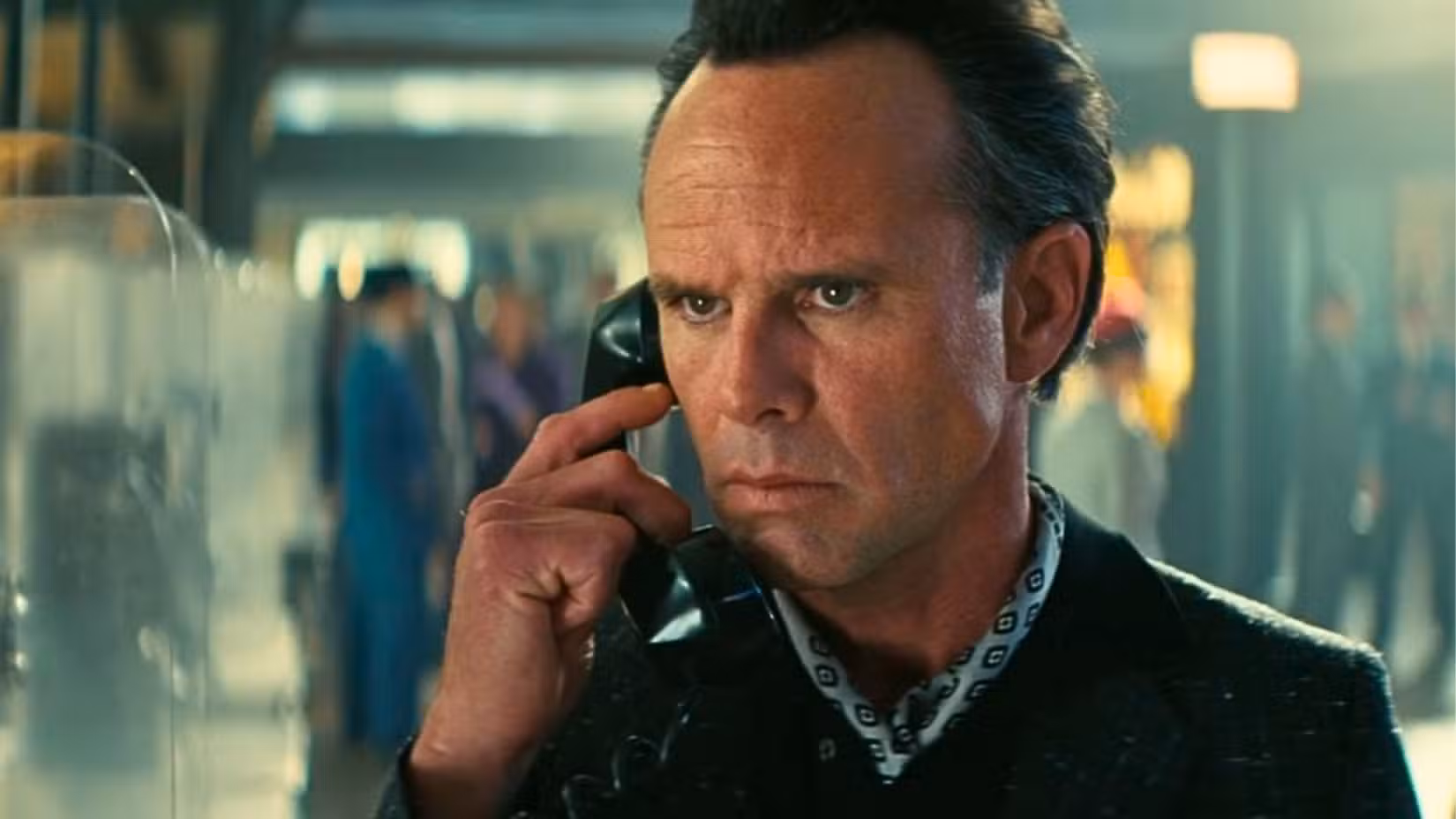 Uma imagem da 2ª temporada de Fallout apresentando Walton Goggins como Cooper Howard. Ele tem cabelo penteado para trás e usa lenço no pescoço. Ele parece preocupado e tem um telefone público encostado no ouvido.