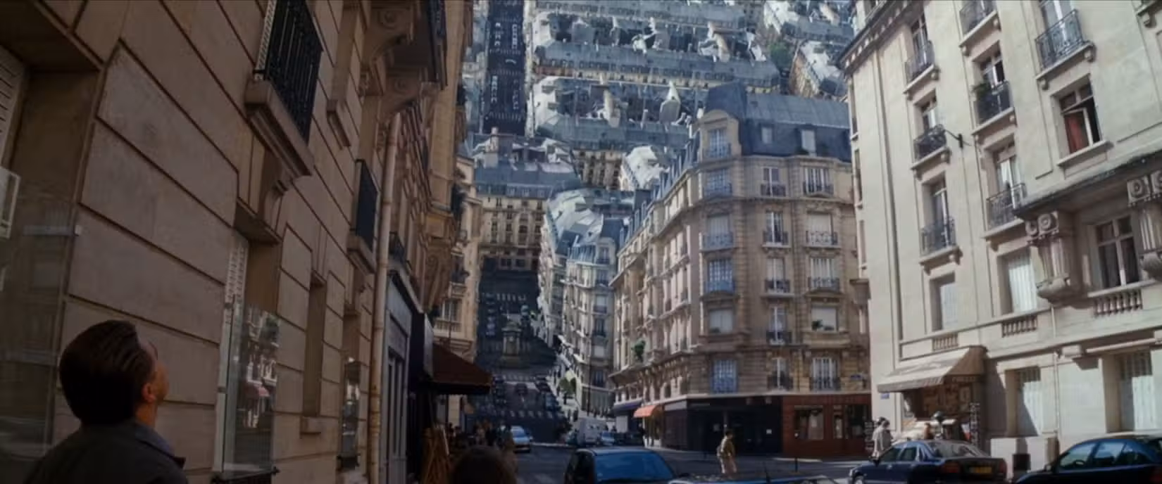 Uma rua da cidade se dobra ao meio, com parte dela aparecendo verticalmente acima de um trecho horizontal, enquanto Dom (Leonardo DiCaprio) observa em uma cena de A Origem, de Christopher Nolan.