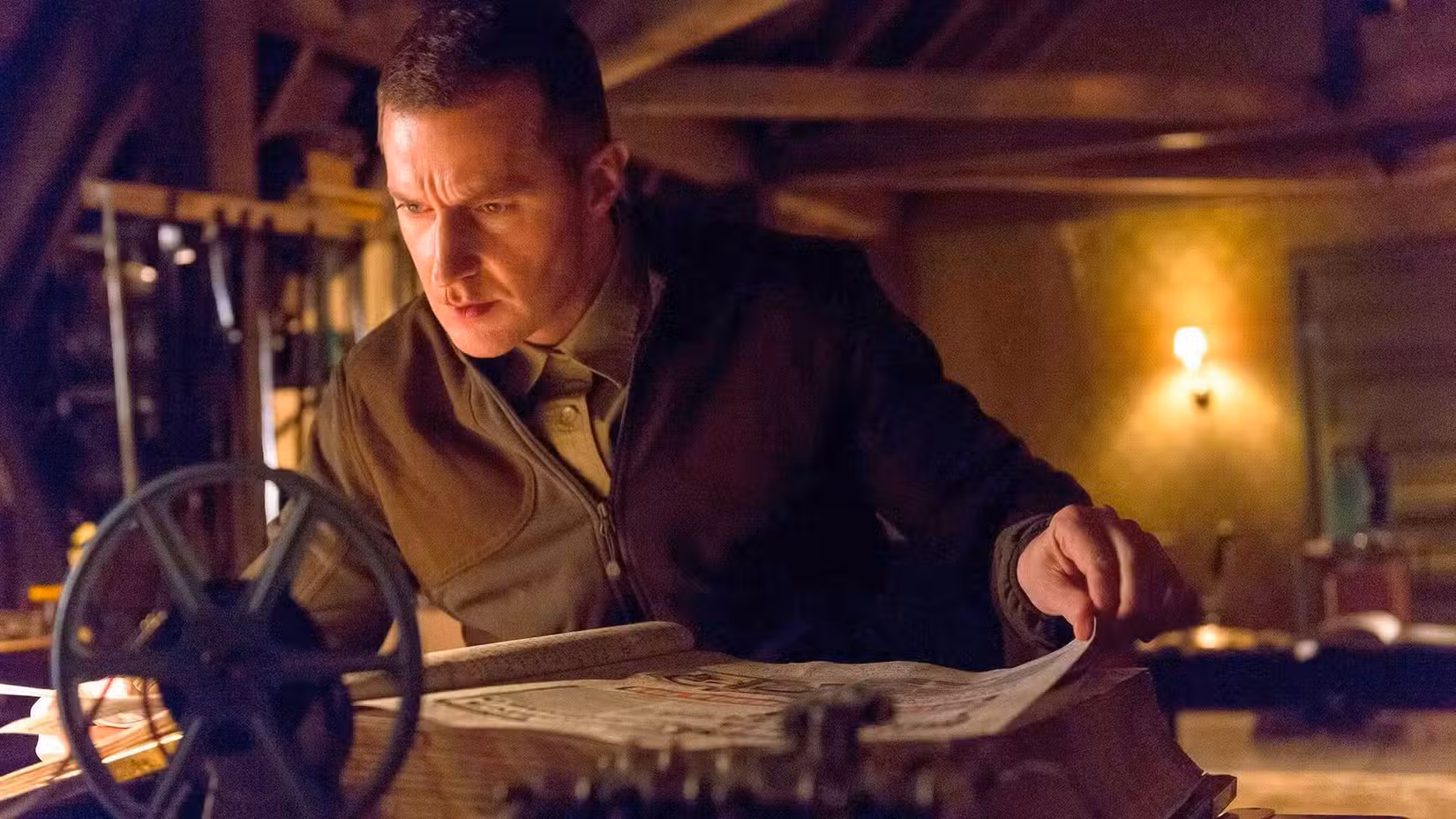 Imagem de Hannibal da NBC. Apresenta Richard Armitage como Francis Dolarhyde. Ele tem cabelo curto e cortado e está olhando por cima de um livro enorme. 