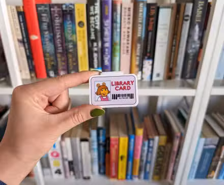 Um pequeno cartão da biblioteca da DW, de Arthur, colocado em frente a uma estante de livros. A mão é de pele clara, com esmalte verde.