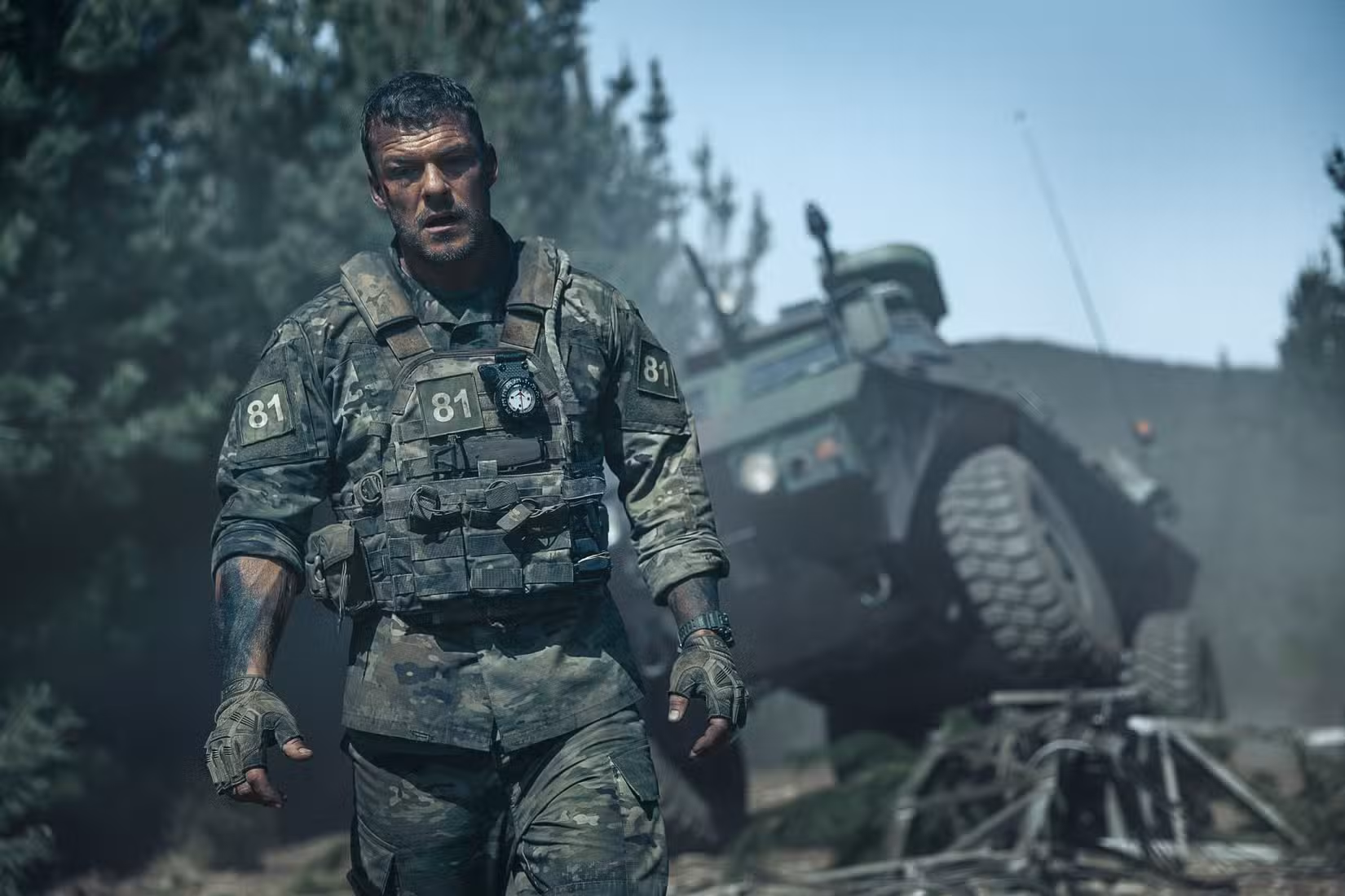 81 (Alan Ritchson), um sargento do Exército com uniforme sujo, coberto de lama e sangue, parado na frente de um veículo militar destruído em War Machine