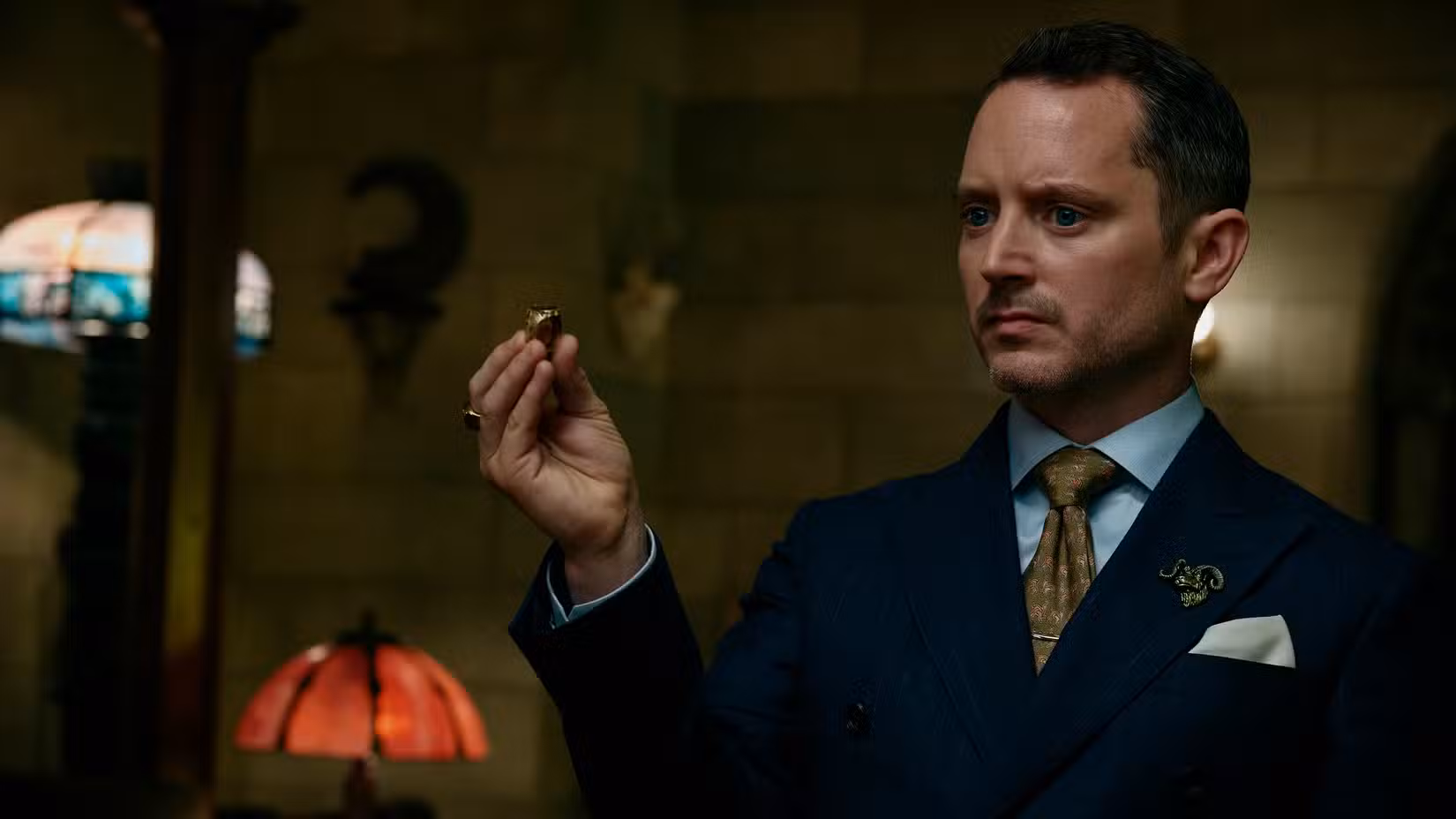 O Advogado (Elijah Wood), um homem vestindo um terno azul com um distintivo de lapela com cabeça de cabra, segura um anel de ouro enquanto está em uma sala iluminada por lâmpadas em Ready or Not 2.