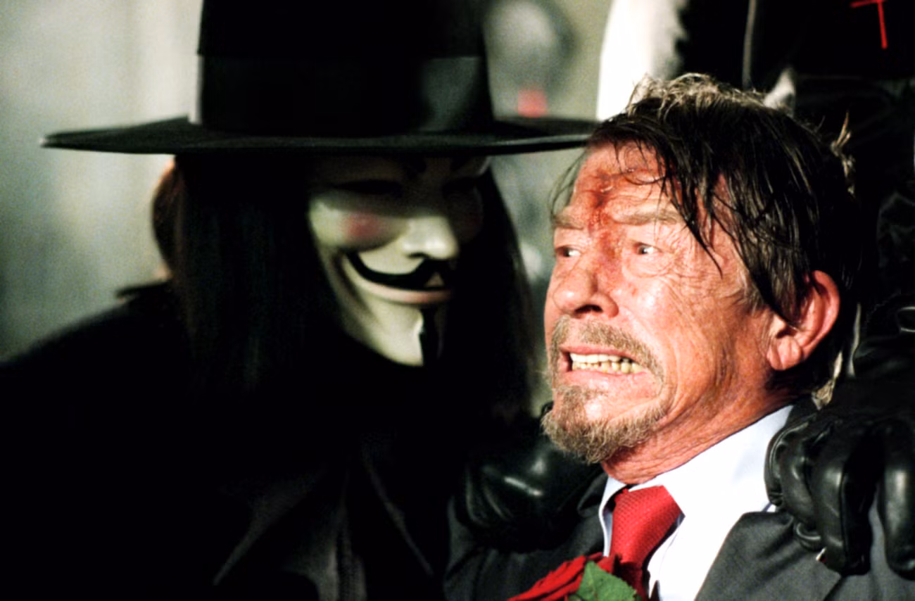 Uma imagem do filme de 2006, "V de Vingança." Destaca John Hurt, ensanguentado, machucado e parecendo assustado, olhando para frente enquanto V (Hugo Weaving) usa uma máscara de Guy Fawkes e se aproxima.