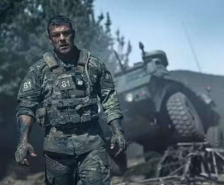 81 (Alan Ritchson), um sargento do Exército com uniforme sujo, coberto de lama e sangue, parado na frente de um veículo militar destruído em War Machine