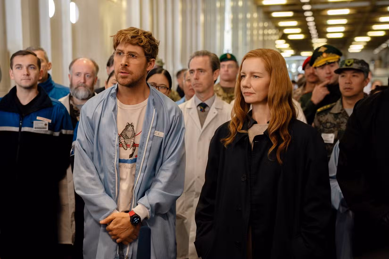 Uma grande multidão de pessoas está aglomerada em um corredor. O cientista Ryland Grace (Ryan Gosling, de óculos e um manto azul solto) e Eva Stratt (Sandra Huller, de jaqueta preta lisa) estão na vanguarda.