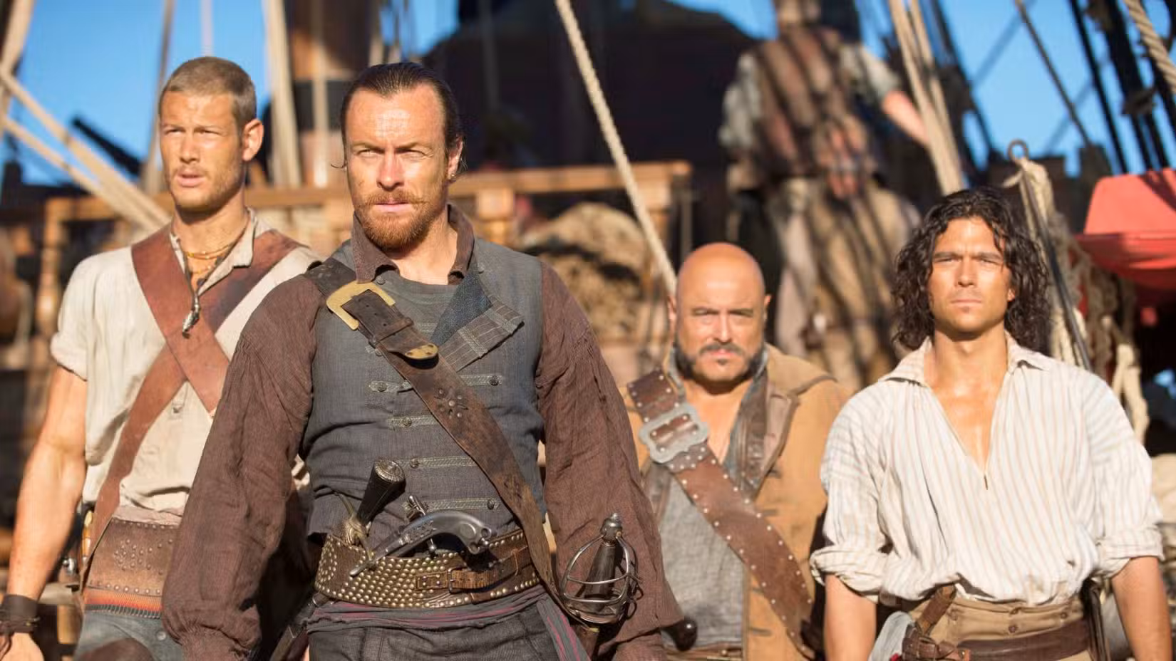 Uma imagem de Black Sails. Mostra Toby Stephens como Capitão James Flint. Ele está flanqueado por três homens.