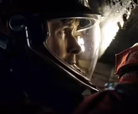 Em um espaço escuro e estreito, o astronauta Ryland Grace (Ryan Gosling), em traje espacial completo e capacete, espia através de uma pequena abertura no Projeto Ave Maria