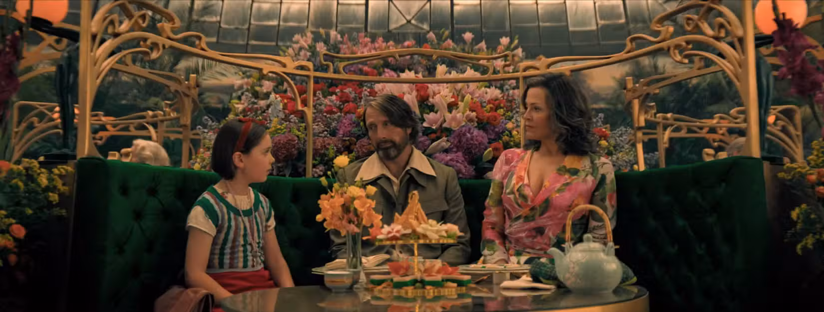 Imagem de Dust Bunny com Sigourney Weaver, Mads Mikkelsen e Sophie Sloan. Os três estão sentados em um café cheio de flores. Um jogo de chá está na frente dos três.