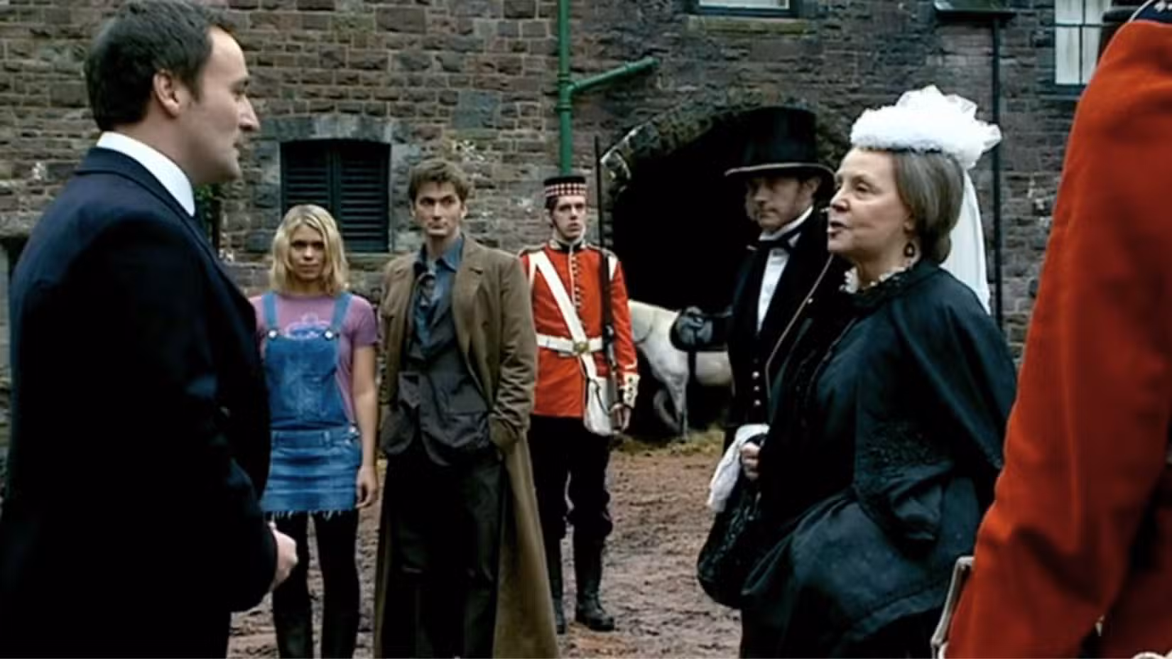 Uma imagem apresentando Billie Piper de Doctor Who e David Tennant como Rose Tyler e o Doutor. A Rainha Vitória, vestida de preto, fala com um homem de terno.