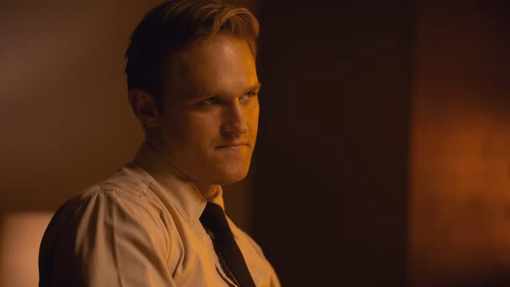 O jovem Lee Shaw (Wyatt Russell), vestindo uma camisa branca e gravata preta, está sentado em um escritório e parece duvidoso em Monarch: Legacy of Monsters, segunda temporada.
