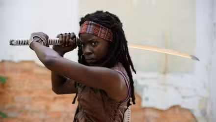 Michonne (Danai Gurira), uma mulher negra com dreadlocks presos por uma bandana colorida, está preparada com uma katana em The Walking Dead
