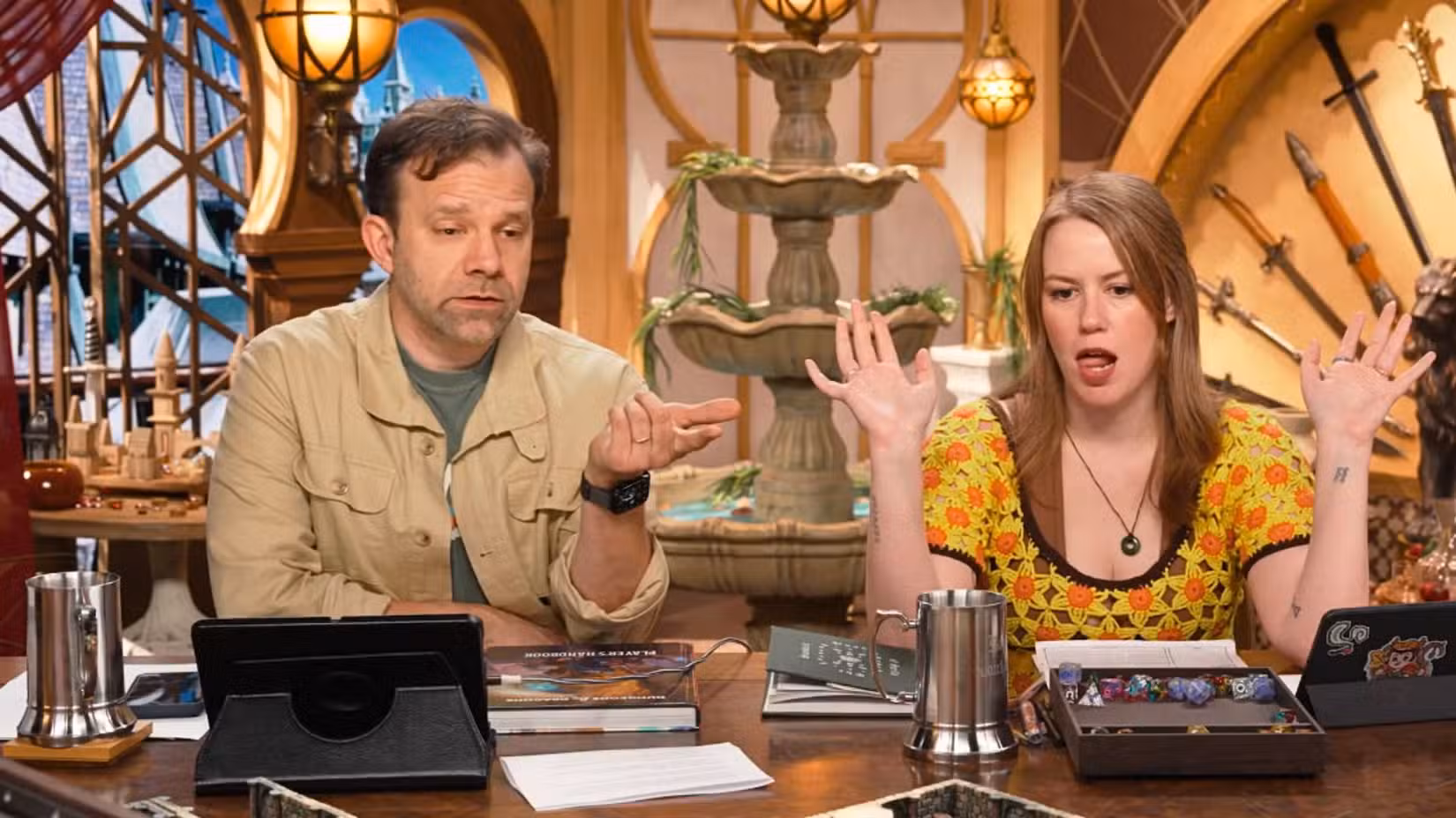 Uma imagem mostrando Liam O'Brien e Marisha Ray da Critical Role sentados lado a lado na mesa da Critical Role.