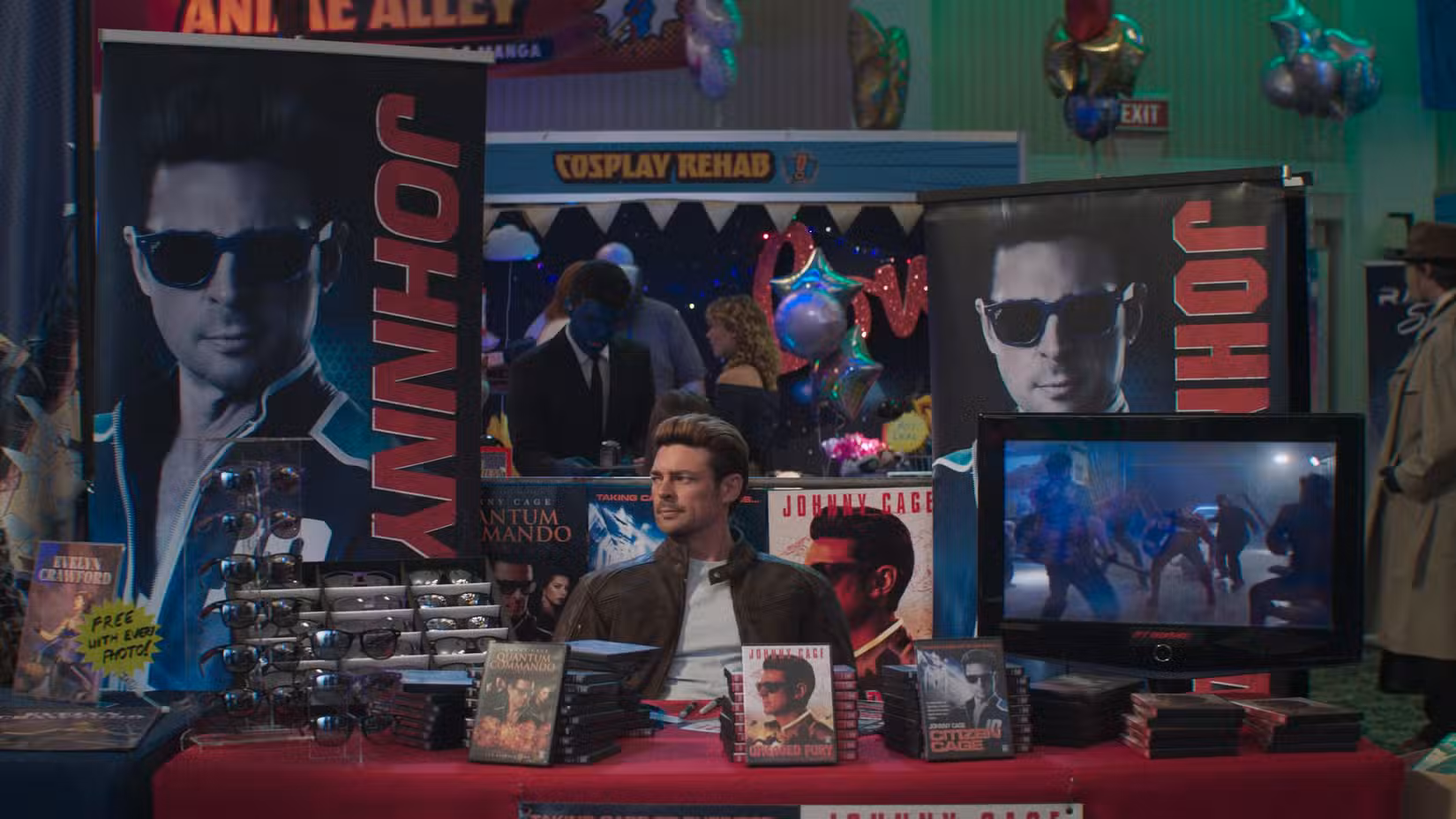 Johnny Cage (Karl Urban) sentado em um estande de uma convenção cercado por DVDs e óculos de sol em Mortal Kombat 2.