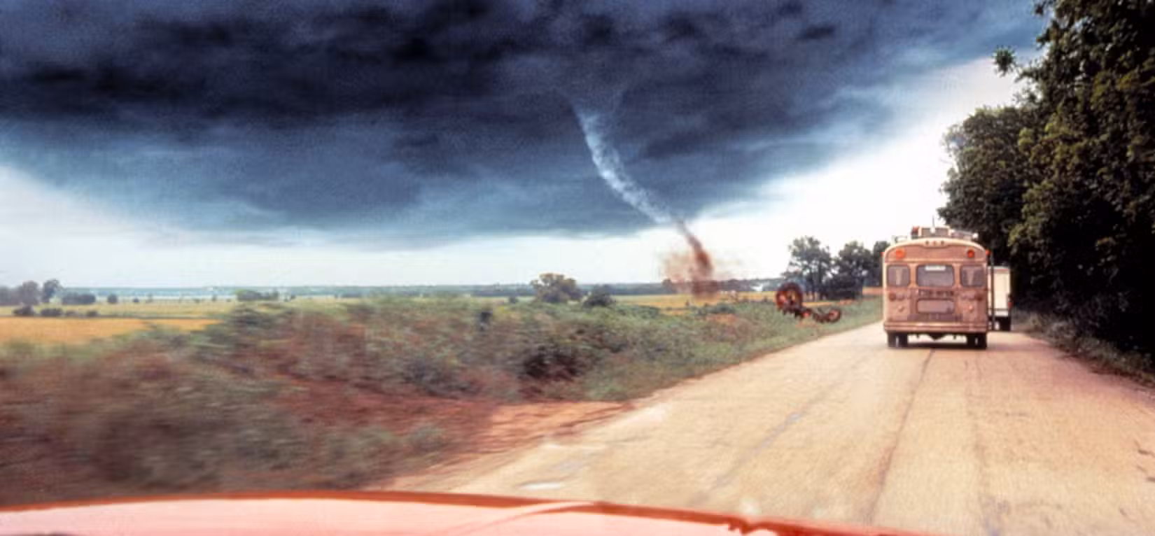 Uma captura em tela ampla mostrando um tornado ao fundo com um ônibus velho visto sobre o capô de um caminhão