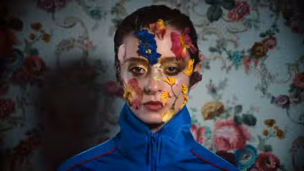 Uma mulher usa um suéter azul em frente a um papel de parede cheio de flores, enquanto flores crescem em seu rosto em Copenhagen Cowboy.